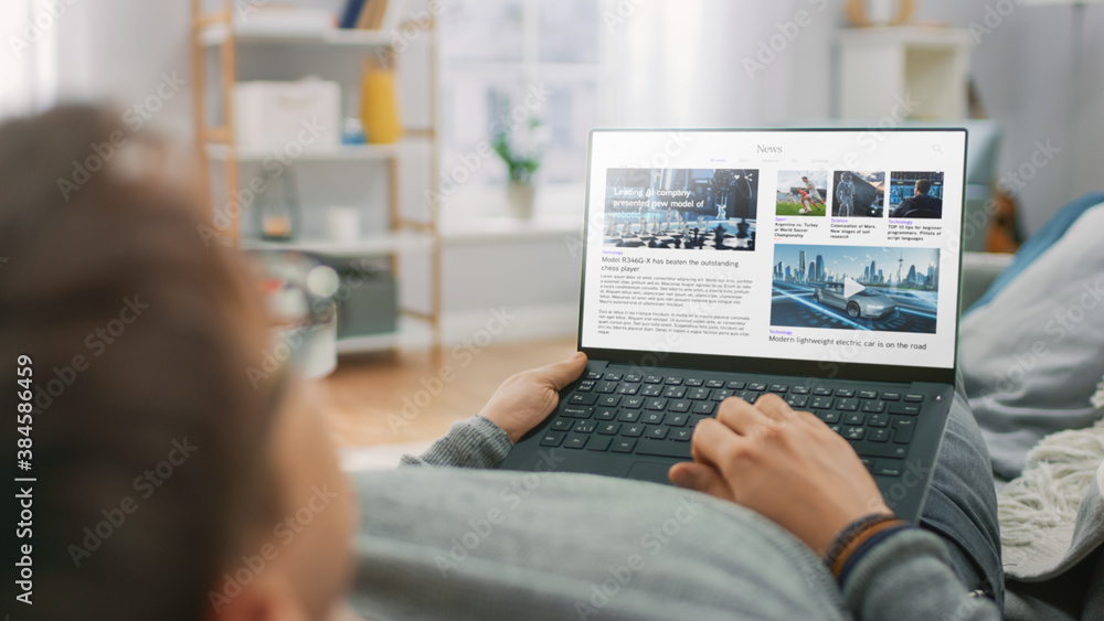 Poster young man at home laying on a couch and using laptop computer for reading news about technological b