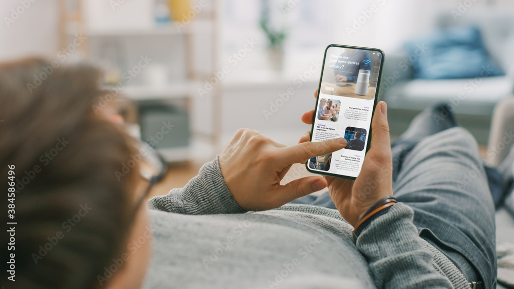 Poster young man at home is lying on a sofa and using smartphone for scrolling and reading news about techn