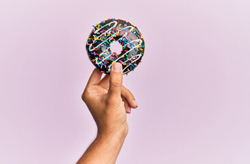 Hand of hispanic man holding chocolate donut over isolated pink background.