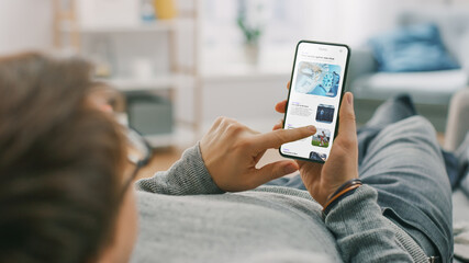 Young Man at Home is Lying on a Coach and Using Smartphone for Scrolling and Reading News about...
