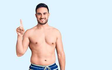 Fototapeta premium Young handsome man wearing swimwear pointing finger up with successful idea. exited and happy. number one.