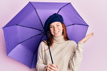 Young red head girl holding purple umbrella wearing fresh beret celebrating victory with happy smile and winner expression with raised hands