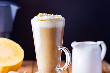 Pumpkin latte and milk jug on wooden boards. Pumpkin spice latte with whipped cream and piece of pumpkin. Hot autumn drink on a burlap