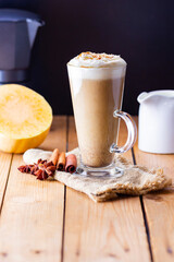 Pumpkin latte and milk jug on wooden boards. Pumpkin spice latte with whipped cream and piece of pumpkin. Hot autumn drink on a burlap