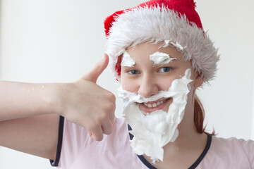 Fototapeta na wymiar A teenage girl in a Santa hat with a beard, mustache and eyebrows made of shaving foam, a close-up of her face, next to her hand with her thumb raised. Preparing for the new year