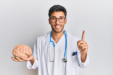 Young handsome man wearing doctor coat holding brain smiling with an idea or question pointing...