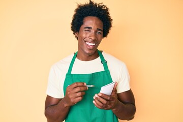 Handsome african american man with afro hair wearing waiter apron taking order winking looking at...