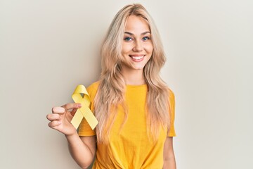 Beautiful caucasian blonde girl holding suicide prevention yellow ribbon looking positive and happy...