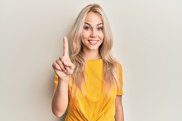 Beautiful caucasian blonde girl wearing casual tshirt showing and pointing up with finger number one while smiling confident and happy.