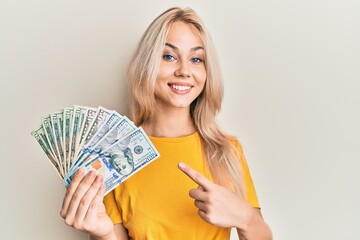 Beautiful caucasian blonde girl holding dollars smiling happy pointing with hand and finger