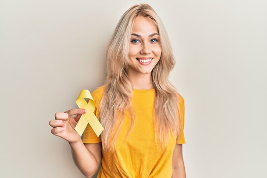 Beautiful caucasian blonde girl holding suicide prevention yellow ribbon looking positive and happy standing and smiling with a confident smile showing teeth