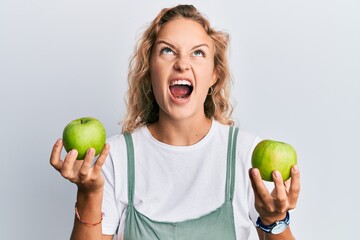 Beautiful caucasian woman holding green apples angry and mad screaming frustrated and furious, shouting with anger looking up.
