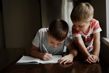 a cute caucasian school boy doing his homework writing letters at the table in kitchen. His juniur brother watching him with interest