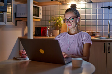 Busy woman student workaholic in headphones preparing for exam, taking notes in notebook during watching webinar and learning online lecture on laptop late in the evening