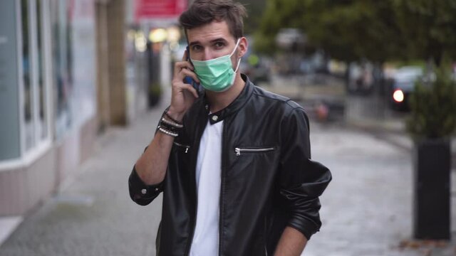 A Young Man With A Cool Hairstyle Wearing A Black Jacket, A White T-shirt And A Green Protective Covid-19 Facemask, Standing On A Busy Street, Picking Up His Phone And Calling, Static Close Up 4k.