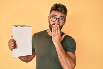 Young hispanic man wearing glasses holding notebook covering mouth with hand, shocked and afraid...