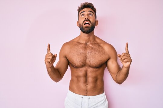 Young hispanic man standing shirtless amazed and surprised looking up and pointing with fingers and raised arms.