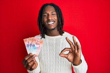 African american man with braids holding 20 swiss franc banknotes doing ok sign with fingers, smiling friendly gesturing excellent symbol