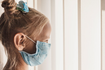 Fototapeta premium Little girl kid in a medical mask by the window with blinds. child on self-isolation, sad, disappointed, hoping for the best. covid backgraund