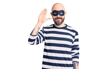 Young handsome man wearing burglar mask waiving saying hello happy and smiling, friendly welcome...
