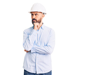 Young handsome man wearing architect hardhat with hand on chin thinking about question, pensive expression. smiling with thoughtful face. doubt concept.