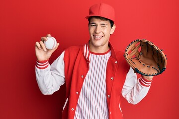 Handsome caucasian man wearing baseball uniform holding golve and ball winking looking at the...