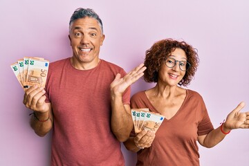 Beautiful middle age couple holding bunch of 50 euro banknotes celebrating achievement with happy smile and winner expression with raised hand