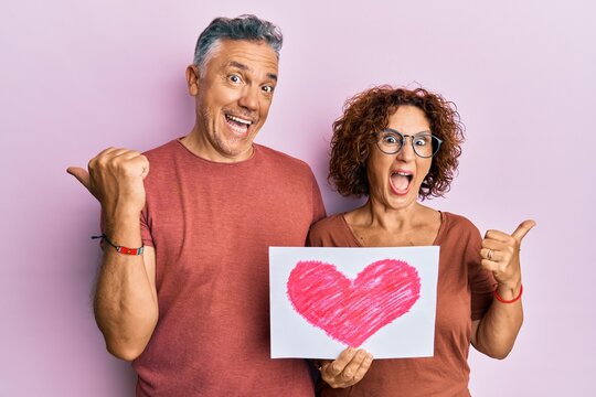 Beautiful middle age couple holding heart draw pointing thumb up to the side smiling happy with open mouth