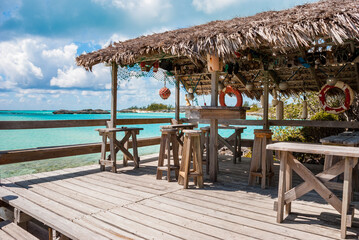 Thatch roof covered wooden patio with tables and chairs overlooking turquoise ocean waters on the Exuma Islands of the Bahamas