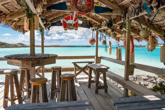 Thatch roof covered wooden patio with tables and chairs overlooking turquoise ocean waters on the Exuma Islands of the Bahamas decorated with buoys, floats, and shoes