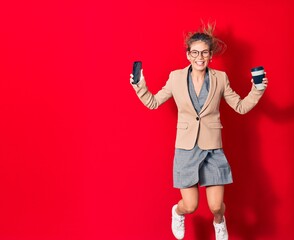 Young beautiful blonde businesswoman wearing elegant clothes and glasses smiling happy. Jumping with smile on face holding smartphone and cup of coffee over isolated red background