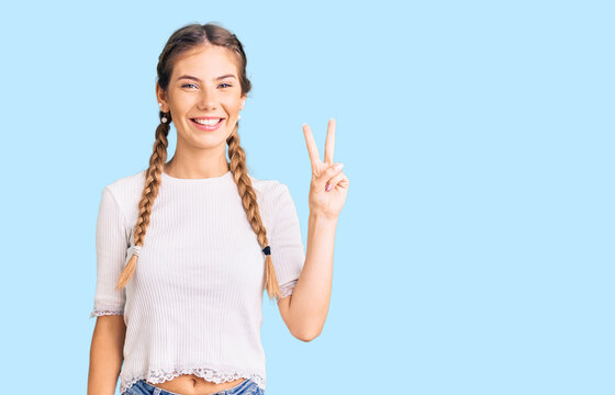 Beautiful caucasian woman with blonde hair wearing braids and white tshirt smiling with happy face winking at the camera doing victory sign. number two.