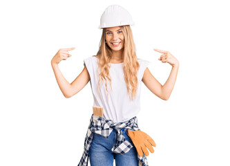 Beautiful caucasian woman with blonde hair wearing hardhat and painter clothes looking confident with smile on face, pointing oneself with fingers proud and happy.