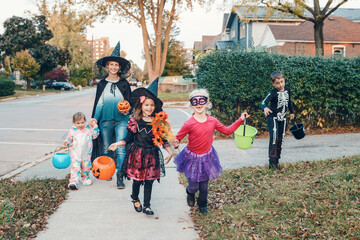 Trick or treat. Mother with children going to trick or treat on Halloween holiday. Mom with kids in party costumes with baskets going to neighbourhood homes for candies, treats. - 384596061
