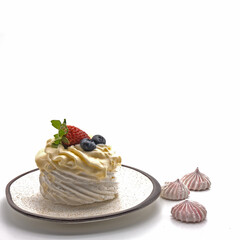 On a white saucer with a rim and dots - meringue, decorated with whipped cream. On top are strawberries, blueberries. Nearby are three tiny pink meringues. Side view. Isolated over white background.