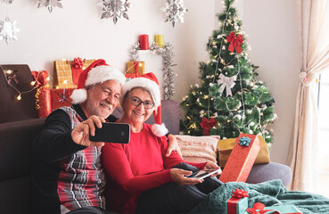 Obraz na płótnie Canvas Happy senior couple with Santa Claus caps enjoying technological devices with Christmas tree and gifts for the family in the background. Love and family concept