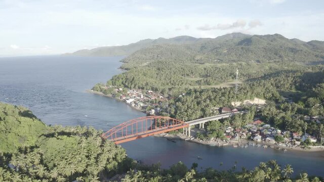Overlooking The Blue Water Surrounded By Forest Mountain Under The Golden Metal Bridge With Vehicles Travelling Near The Small Fishing Village In A Province Of Leyte, Philippines. - Aerial Drone Shot