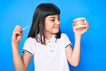 Young little girl with bang holding invisible aligner orthodontic and braces smiling looking to the side and staring away thinking.