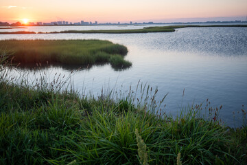 The sun peeks over the horizon in a salt marsh with highrises of Ocean City, Maryland in the...