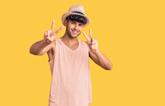 Young hispanic man wearing summer hat smiling looking to the camera showing fingers doing victory sign. number two.
