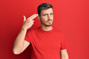 Fototapeta premium Handsome caucasian man wearing casual red tshirt shooting and killing oneself pointing hand and fingers to head like gun, suicide gesture.
