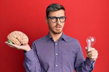 Handsome caucasian man holding brain and lightbulb for inspiration and idea clueless and confused...