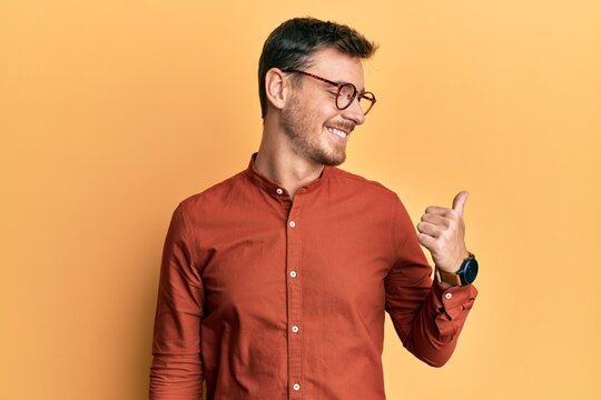 Handsome caucasian man wearing casual clothes and glasses pointing thumb up to the side smiling happy with open mouth