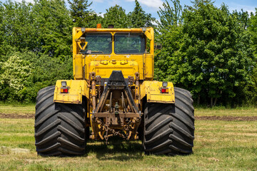 Backside of a yellow tractor with heavy wheels in a field with trees in background