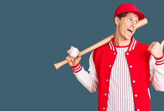 Young handsome man playing baseball holding bat and ball pointing thumb up to the side smiling happy with open mouth