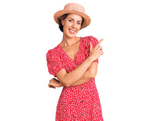 Young beautiful woman wearing summer hat cheerful with a smile of face pointing with hand and finger up to the side with happy and natural expression on face