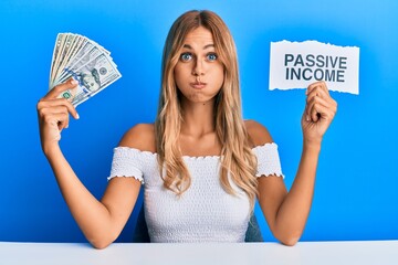 Beautiful blonde young woman holding dollars and passive income text puffing cheeks with funny...