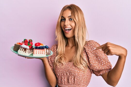 Beautiful blonde young woman holding cake slices pointing finger to one self smiling happy and proud