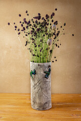 Autumn still life with Sanguisorba officinalis and poppy heads in handmade ceramic vase with green dragonflies placed on wooden board in front of brown wall with clay plaster