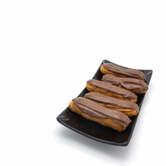 Five fresh eclairs with chocolate icing are on a black rectangular plate. Angle view. Isolated over white background.
